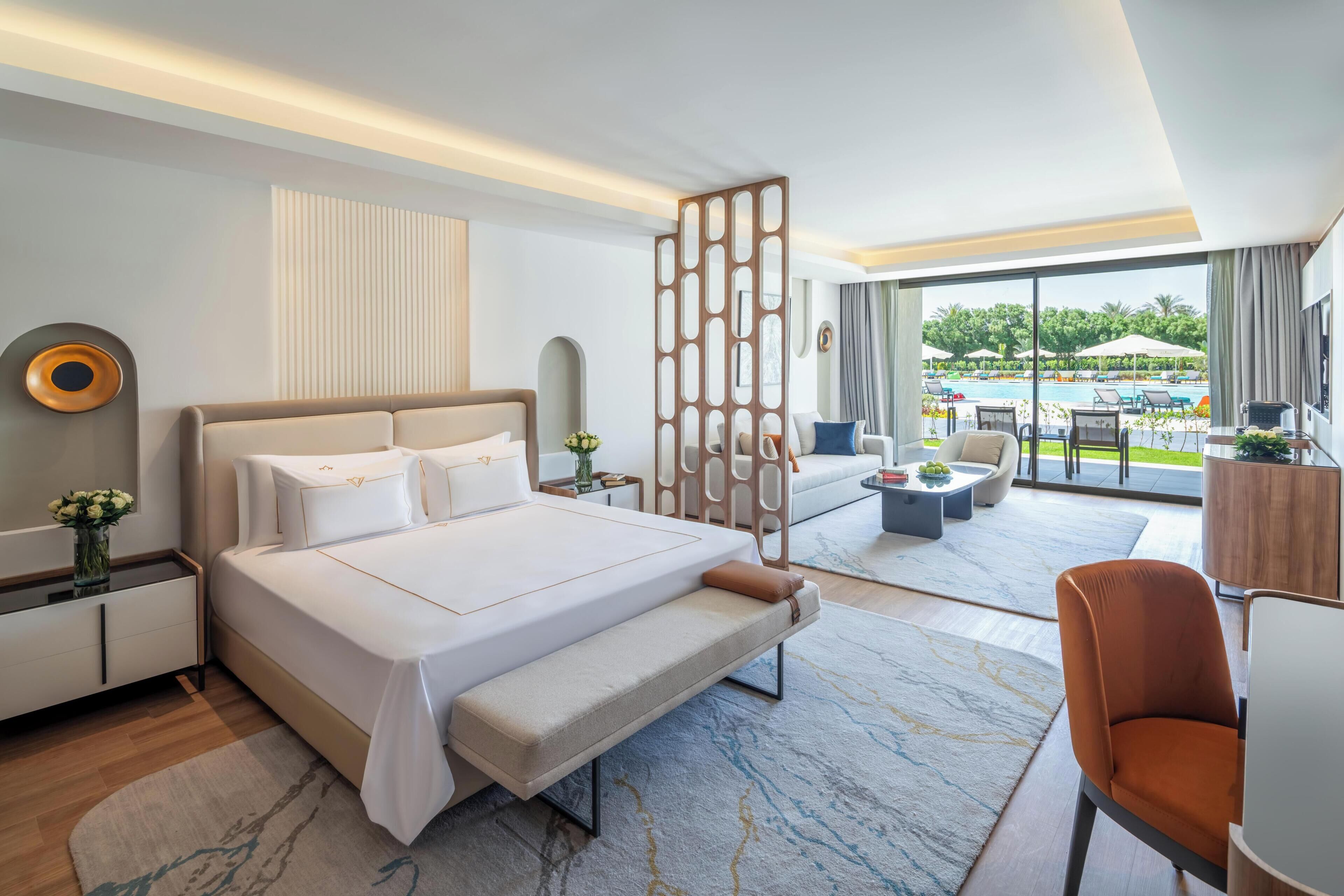 A modern hotel room with a large bed, a sofa, a chair, a desk, and a view of an outdoor pool.