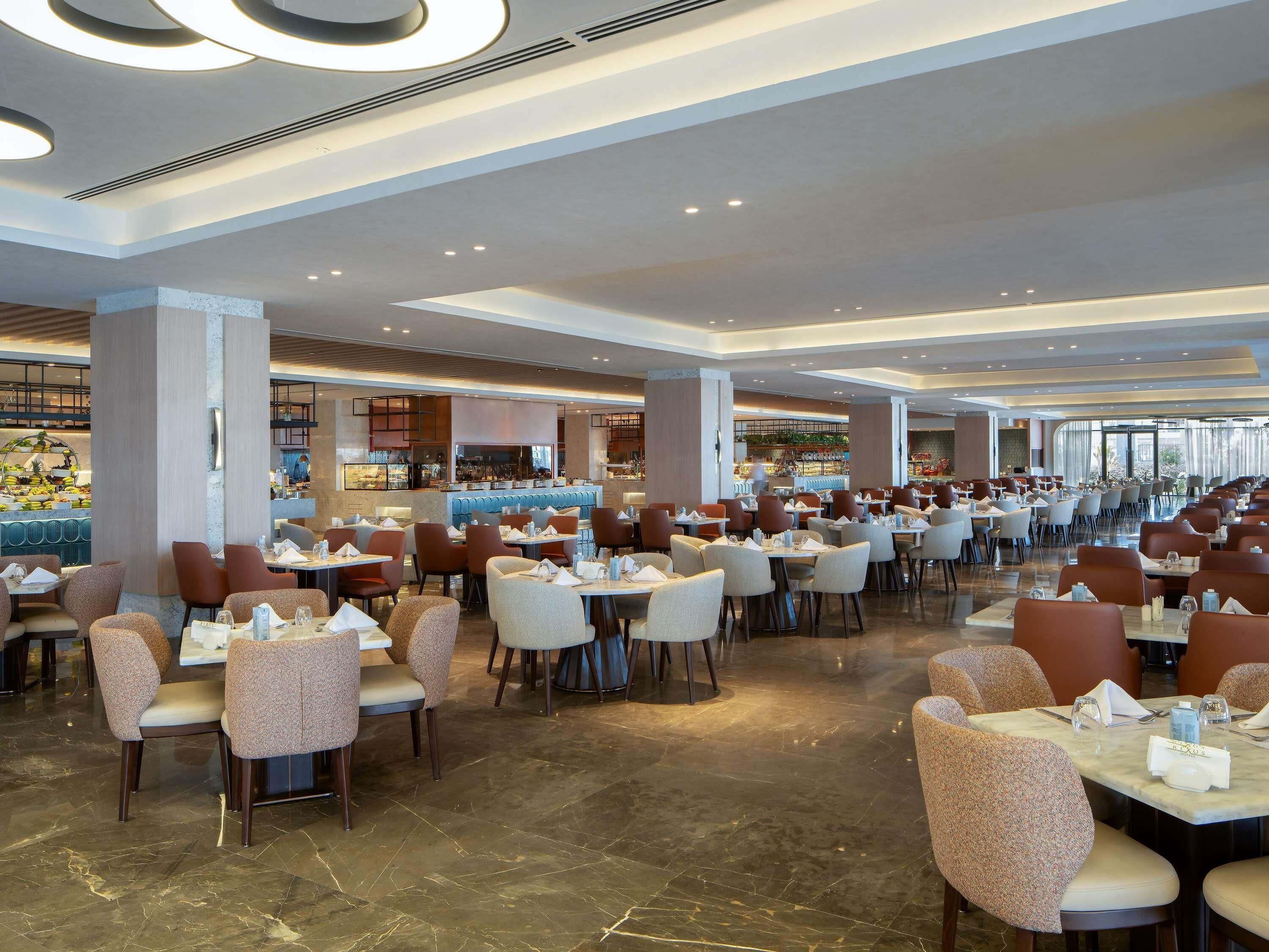 A spacious restaurant with multiple dining tables and chairs, a bar area, and a view of the cityscape through large windows.