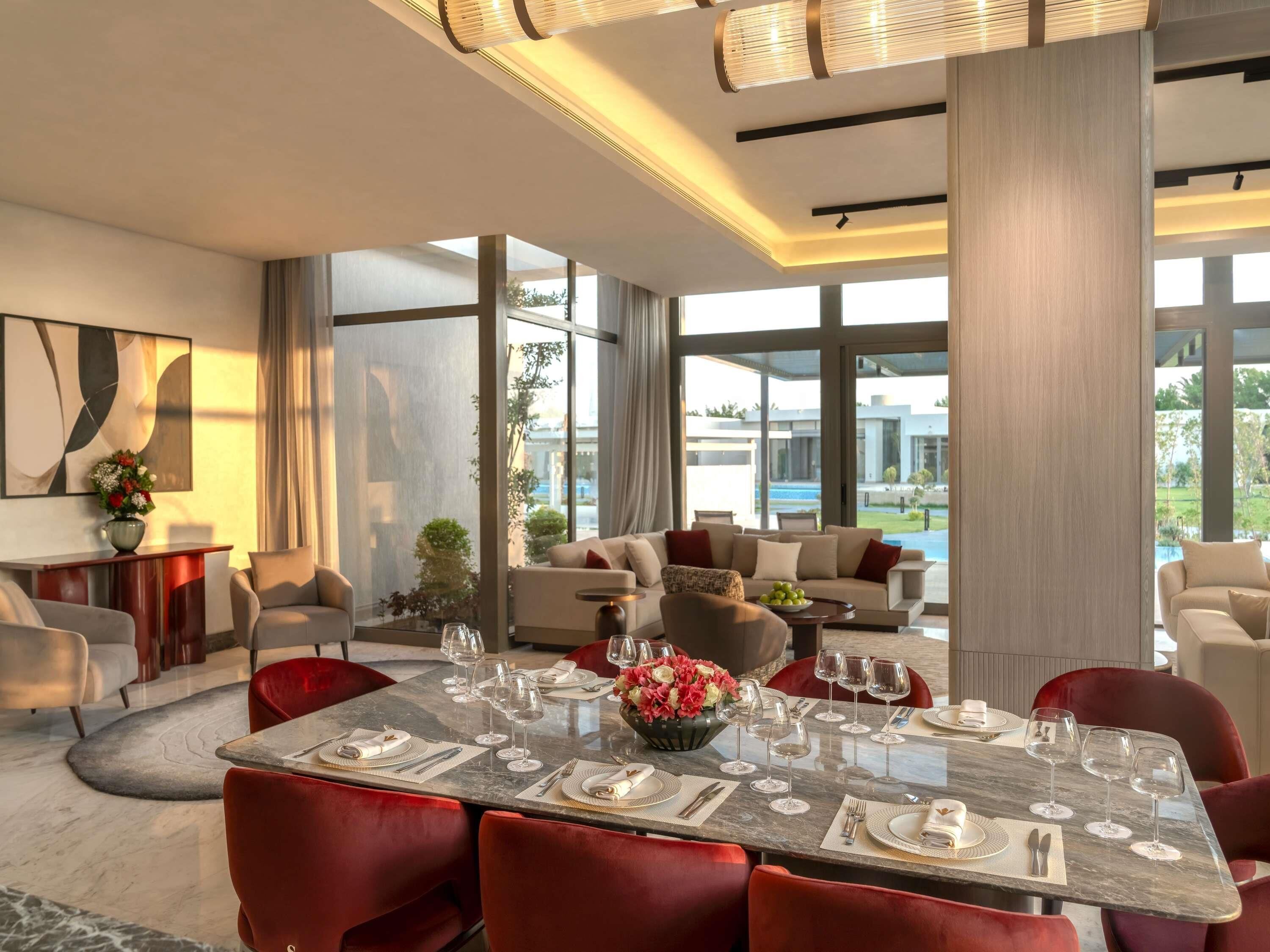 A modern dining area with a marble table, red upholstered chairs, and a view of a pool and greenery outside.