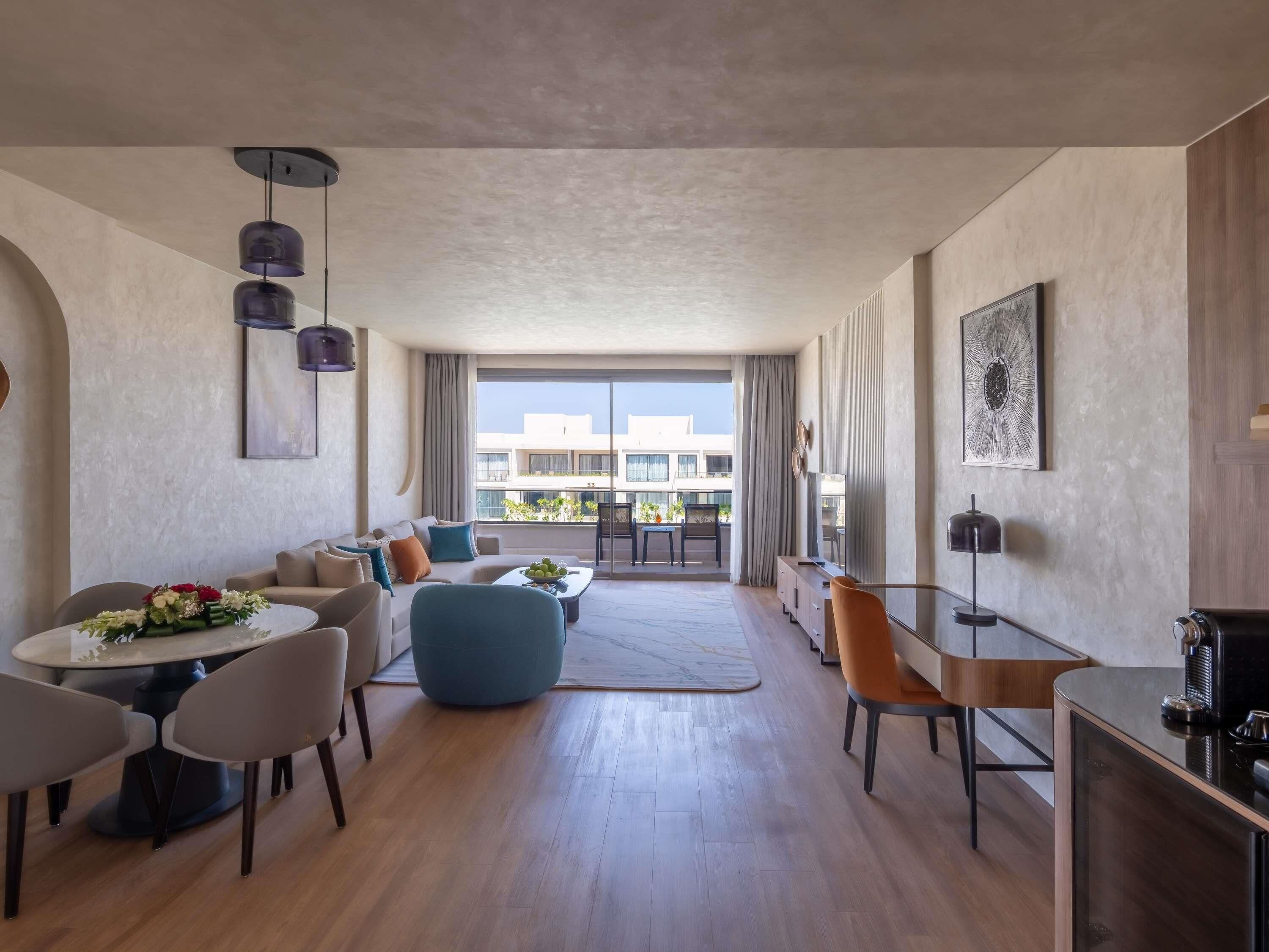 A modern living room with a sofa, coffee table, and chairs, a dining area with a table and chairs, a kitchen area with a counter and appliances, and large windows with a view of buildings.