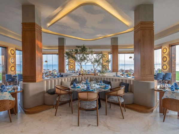 A restaurant with a curved seating arrangement, large windows with ocean views, and a ceiling with a wave design. - Rixos Premium Magawish Bay View (Hurghada)