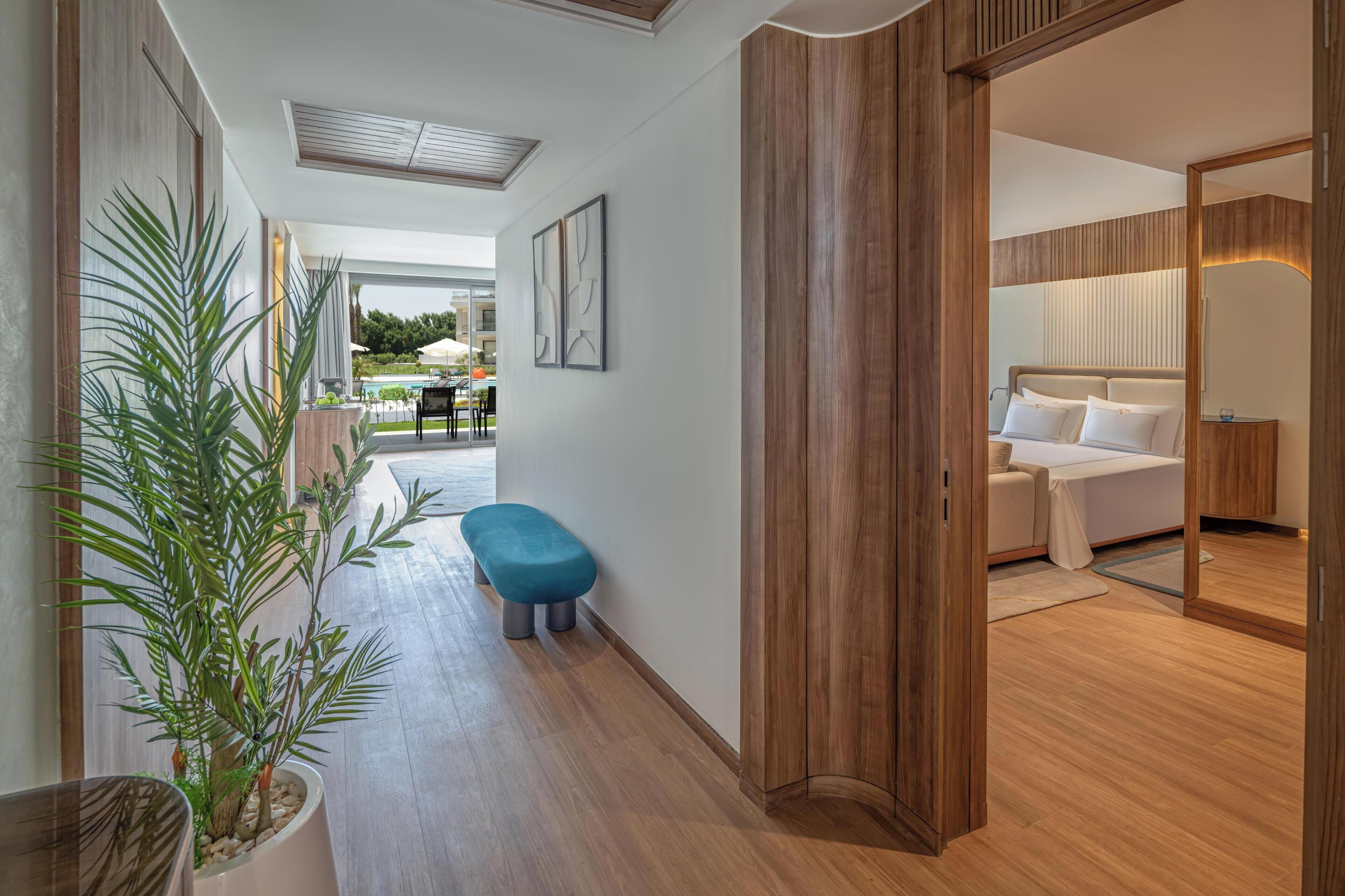 A modern hotel room with a wooden floor, a bench, a bed with white linens, and a large window.