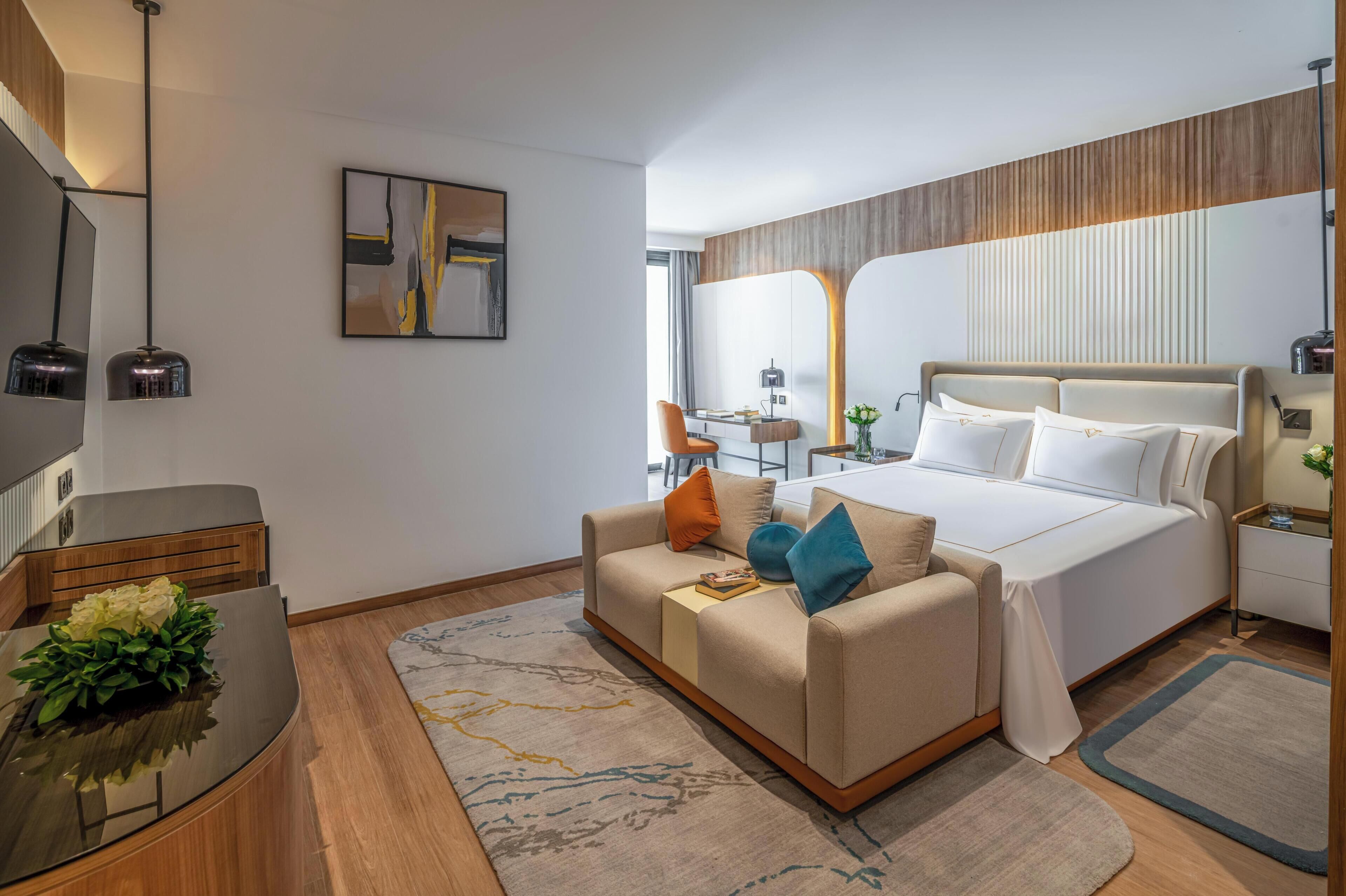 Quarto de hotel moderno com cama, sofá, escrivaninha, espelho e televisão.