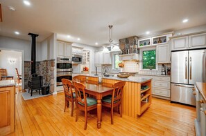 A modern kitchen with white cabinets, a central island, and a stone fireplace. - New! Dog Friendly Lake Access Home w/ Hot Tub (Swanton)