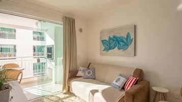 Un salon avec un canapé, une petite table, une plante en pot et un balcon donnant sur des immeubles.