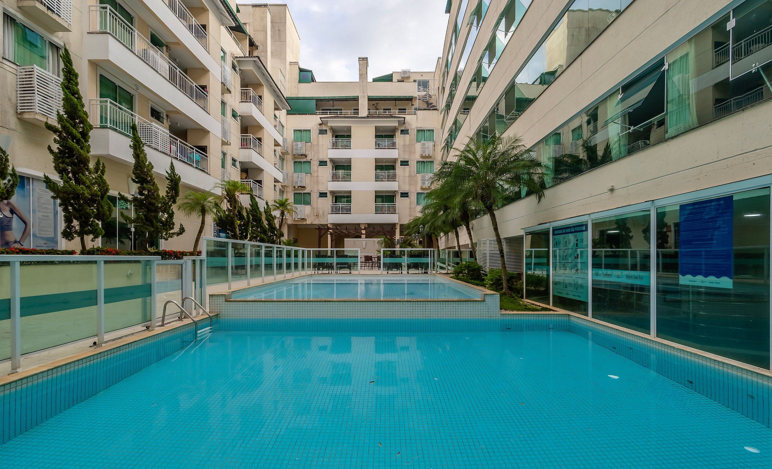 Piscina cercada por um edifício com vários andares, varandas e fachada de vidro.