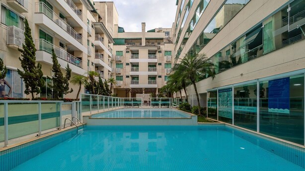 A swimming pool surrounded by a multi-story building with balconies and a glass facade. - Beautiful apt w/ Leisure Facilities Sbh0237 (Bombinhas)
