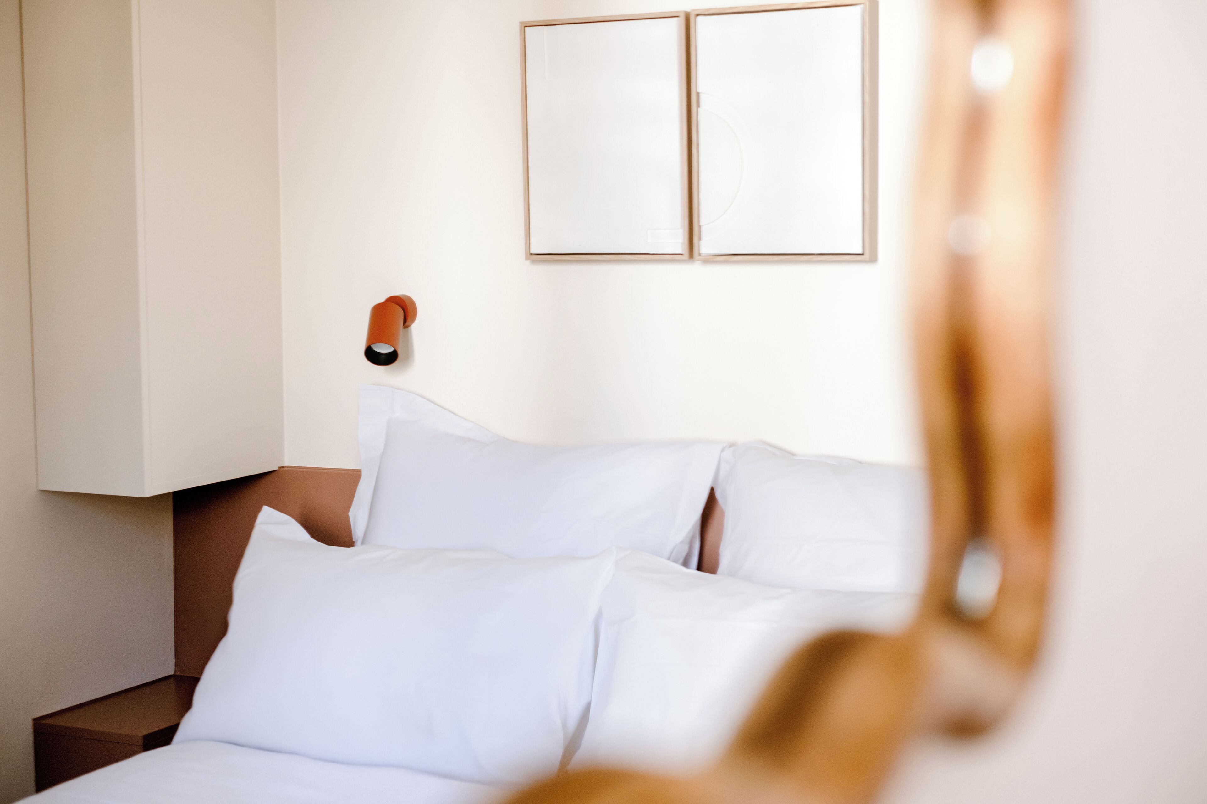 A hotel room with a bed, pillows, a nightstand, and a wall-mounted lamp.
