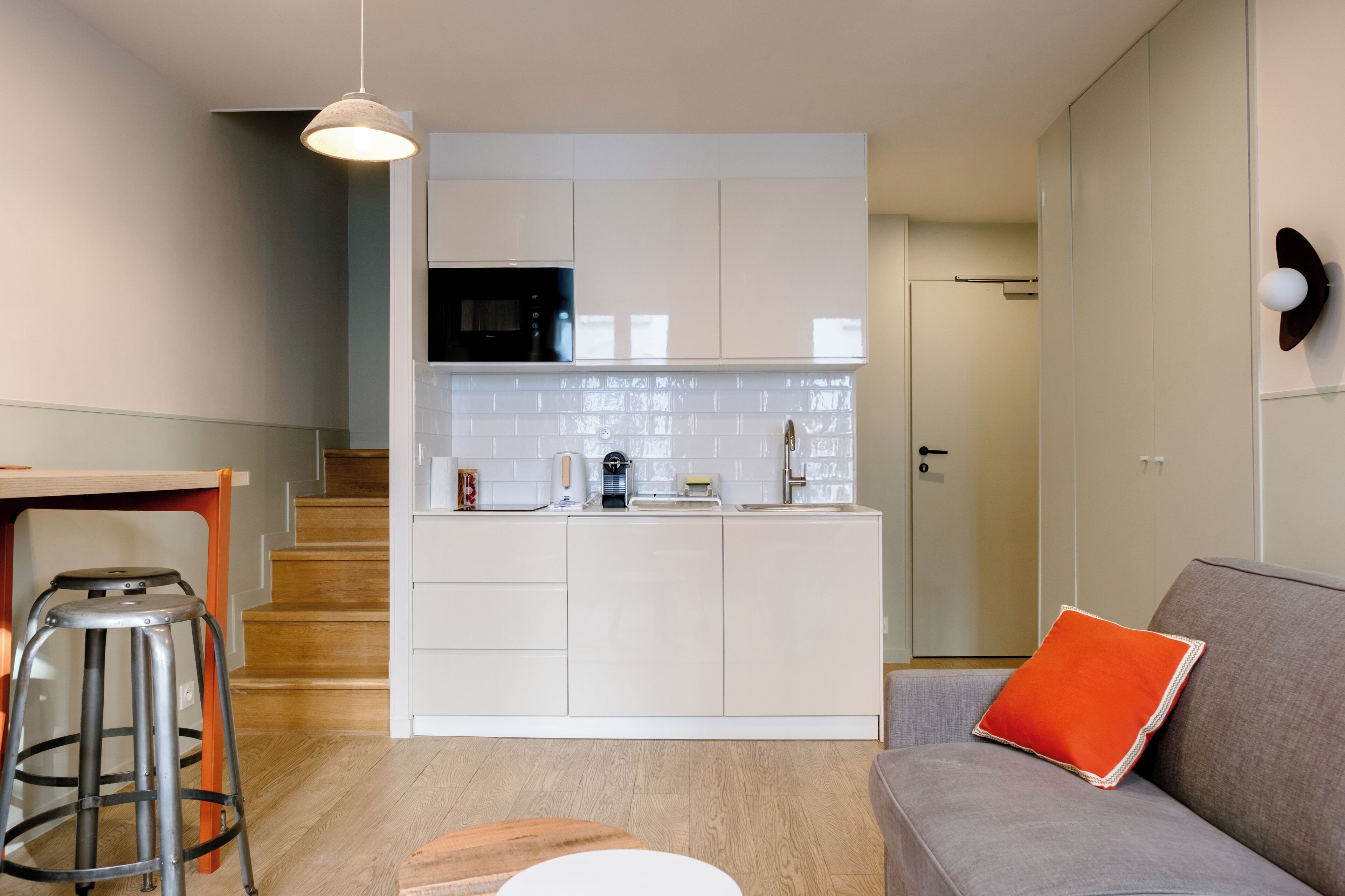 A modern kitchen with white cabinets, a black microwave, and a coffee machine. There is a wooden staircase leading to another room.