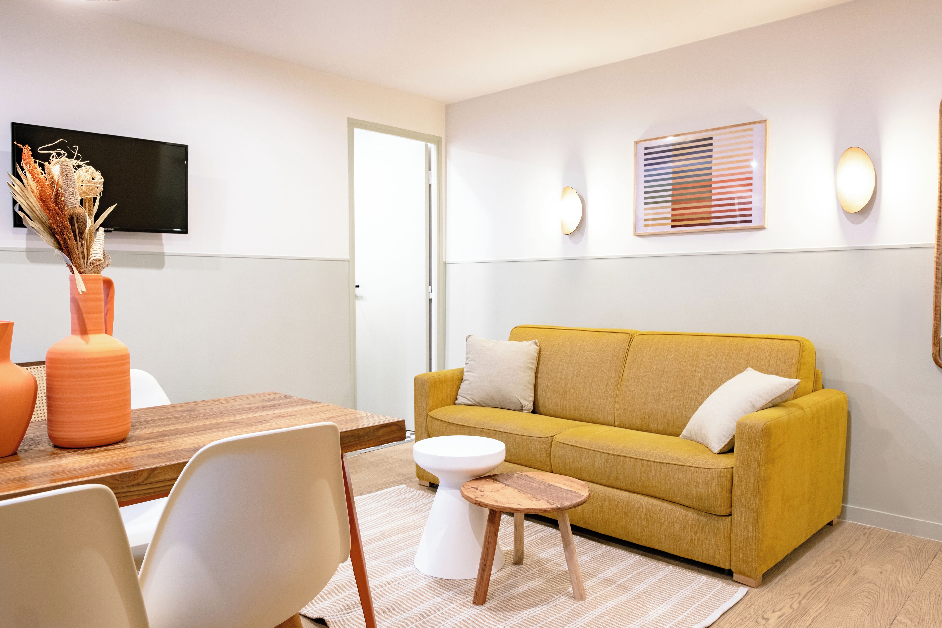 A modern living room with a yellow sofa, white chairs, a wooden coffee table, and a flat-screen TV mounted on the wall.