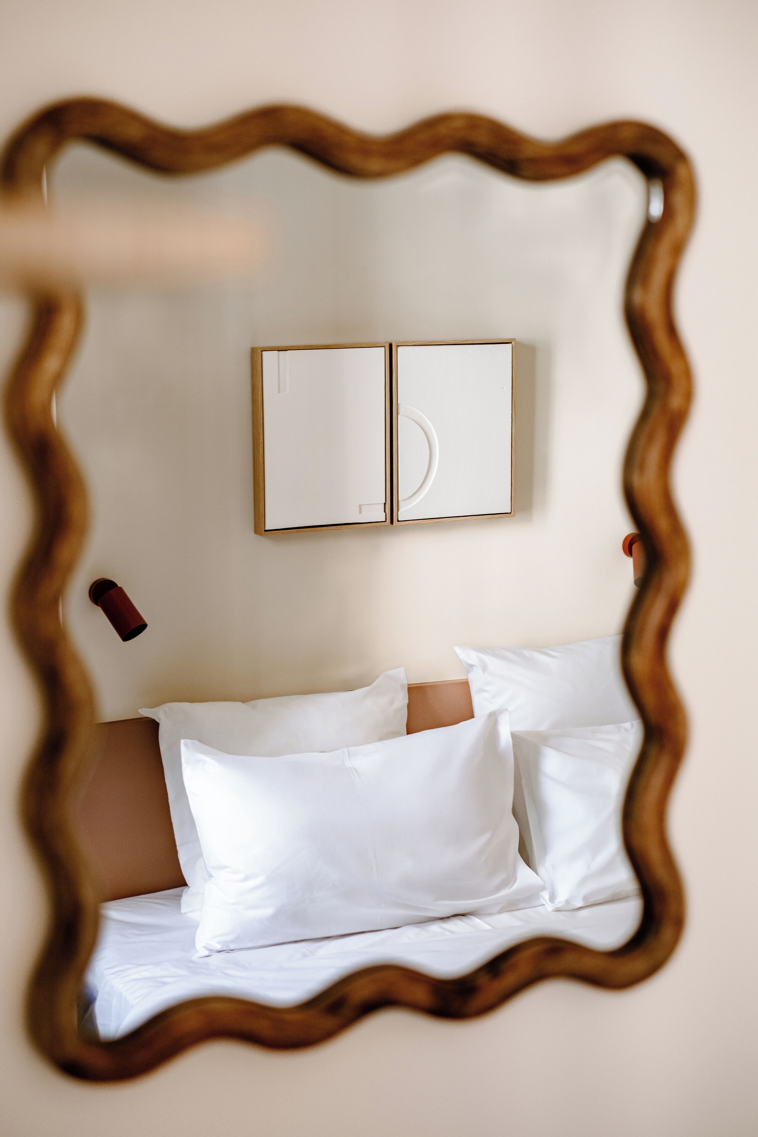 Ein Schlafzimmer mit einem Bett, weißen Kissen und einem spiegelnden, holzrahmenartigen Spiegel, der den Raum zeigt.