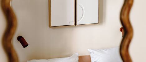 Ein Schlafzimmer mit einem Bett, weißen Kissen und einem spiegelnden, holzrahmenartigen Spiegel, der den Raum zeigt.