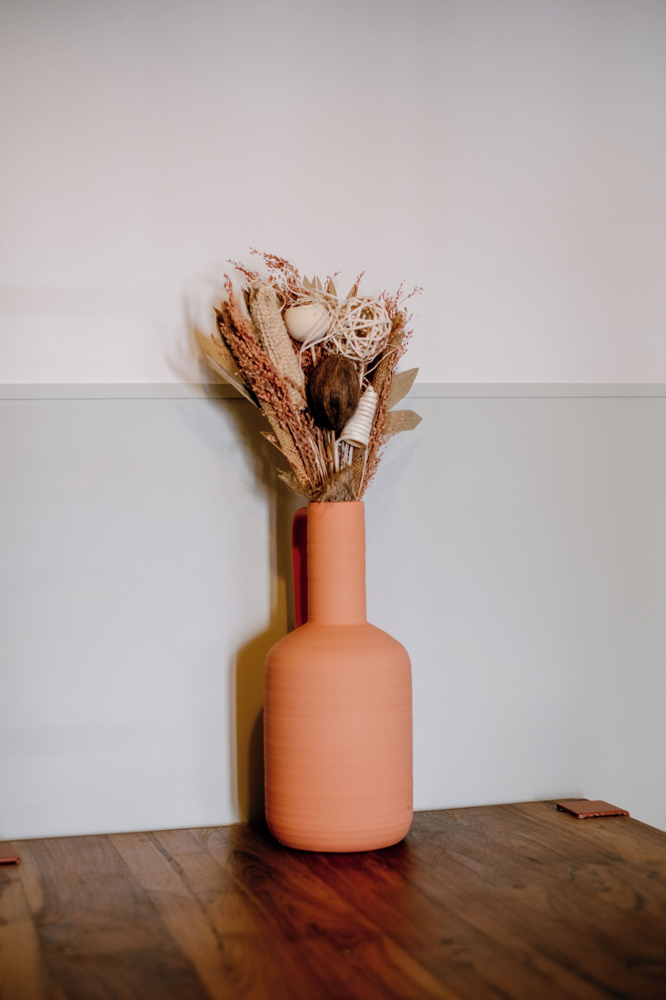 Un vase contenant des fleurs séchées, posé sur une table en bois devant un mur uni.