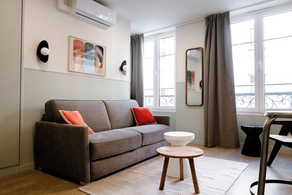 A modern living room with a grey sofa, a round wooden side table, a white stool, and a large window with curtains. - Babylon Popincourt (Paris)