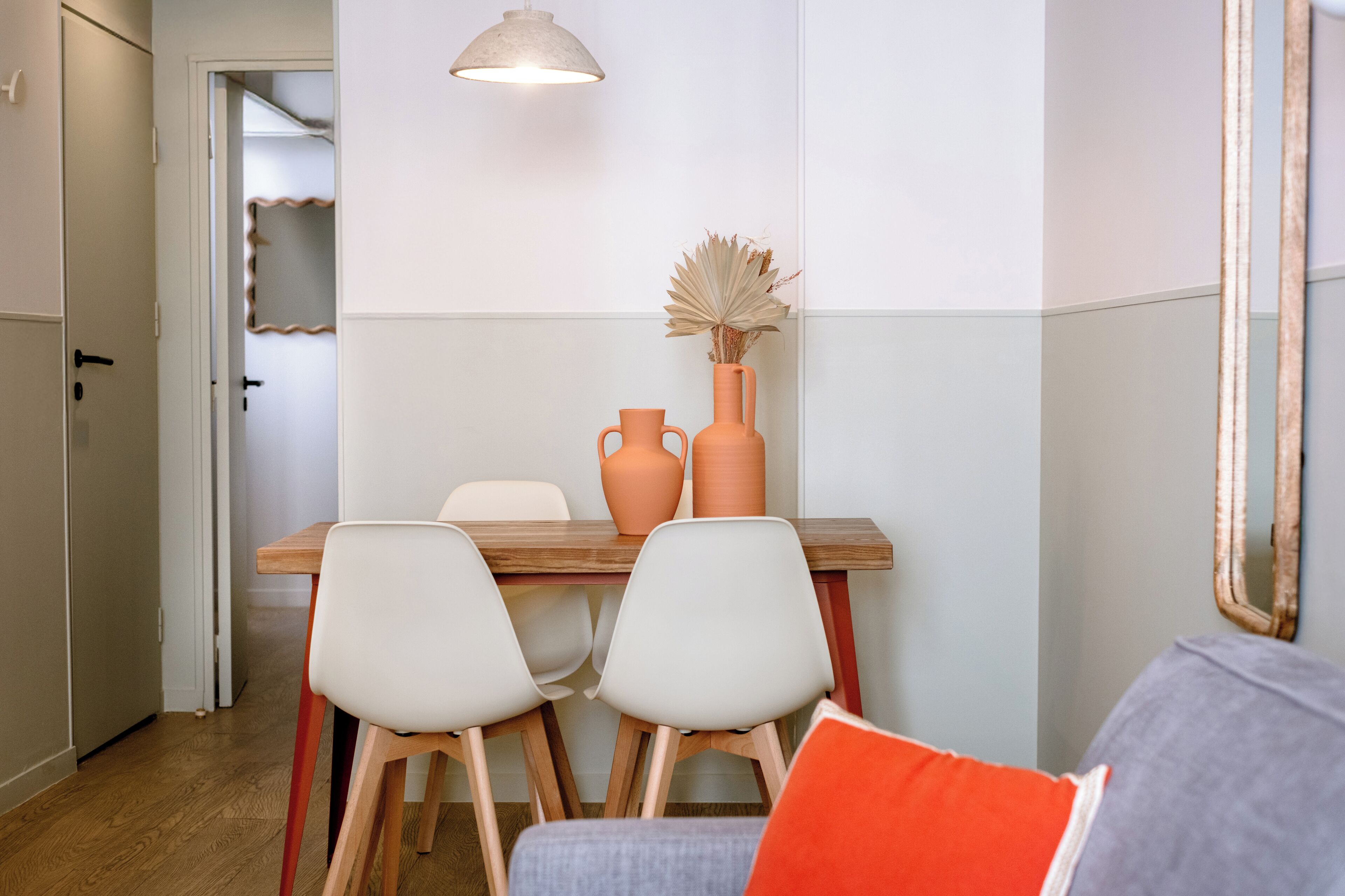 A dining area with a wooden table, two white chairs, a vase with dried plants, and a pendant light.