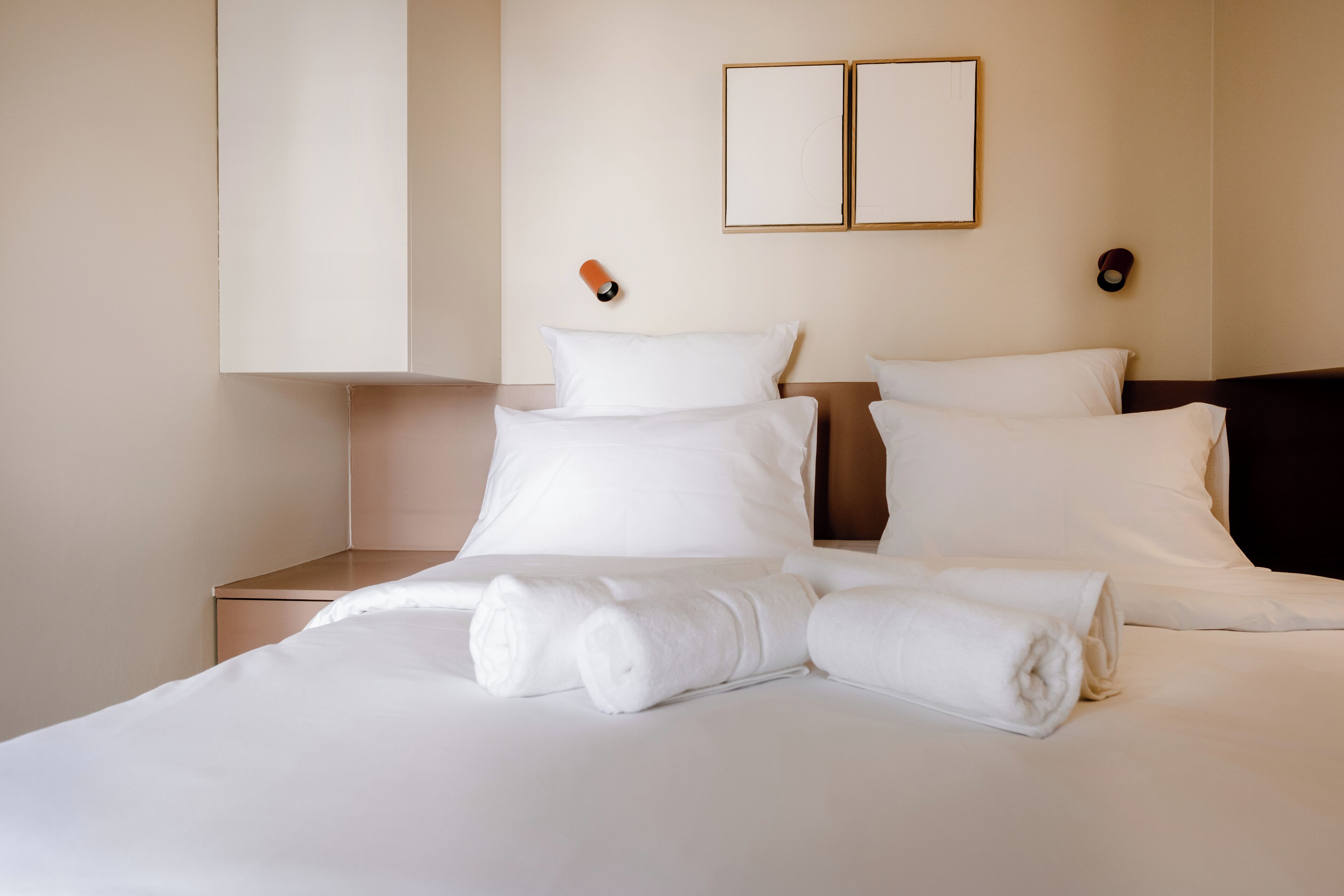 Ein ordentlich gemachtes Bett mit weißen Leinen und aufgerollten Handtüchern, ein Nachttisch und zwei Wandleuchten.