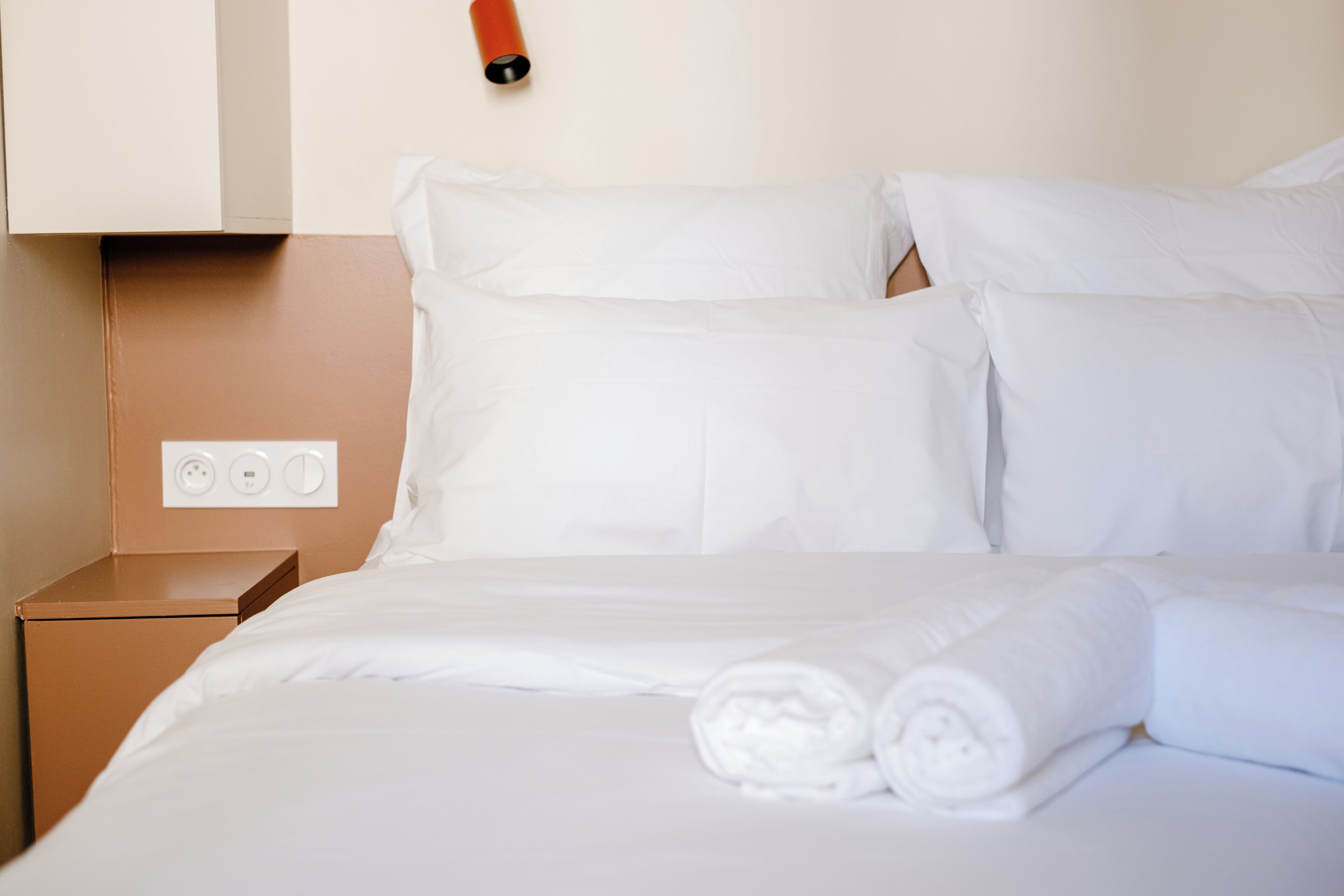 A neatly made bed with white linens and rolled towels, a brown nightstand with a power outlet, and a red object on the wall.
