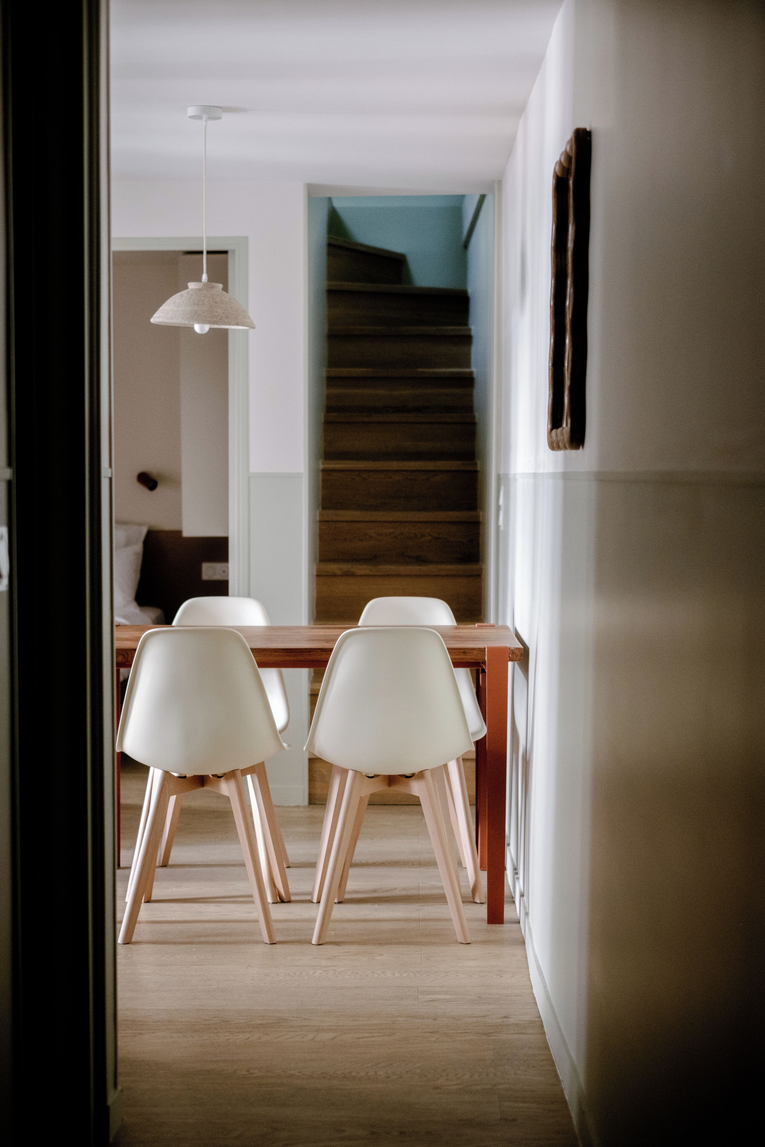 Un coin repas avec une table en bois et des chaises blanches, un luminaire suspendu et un escalier en arrière-plan.