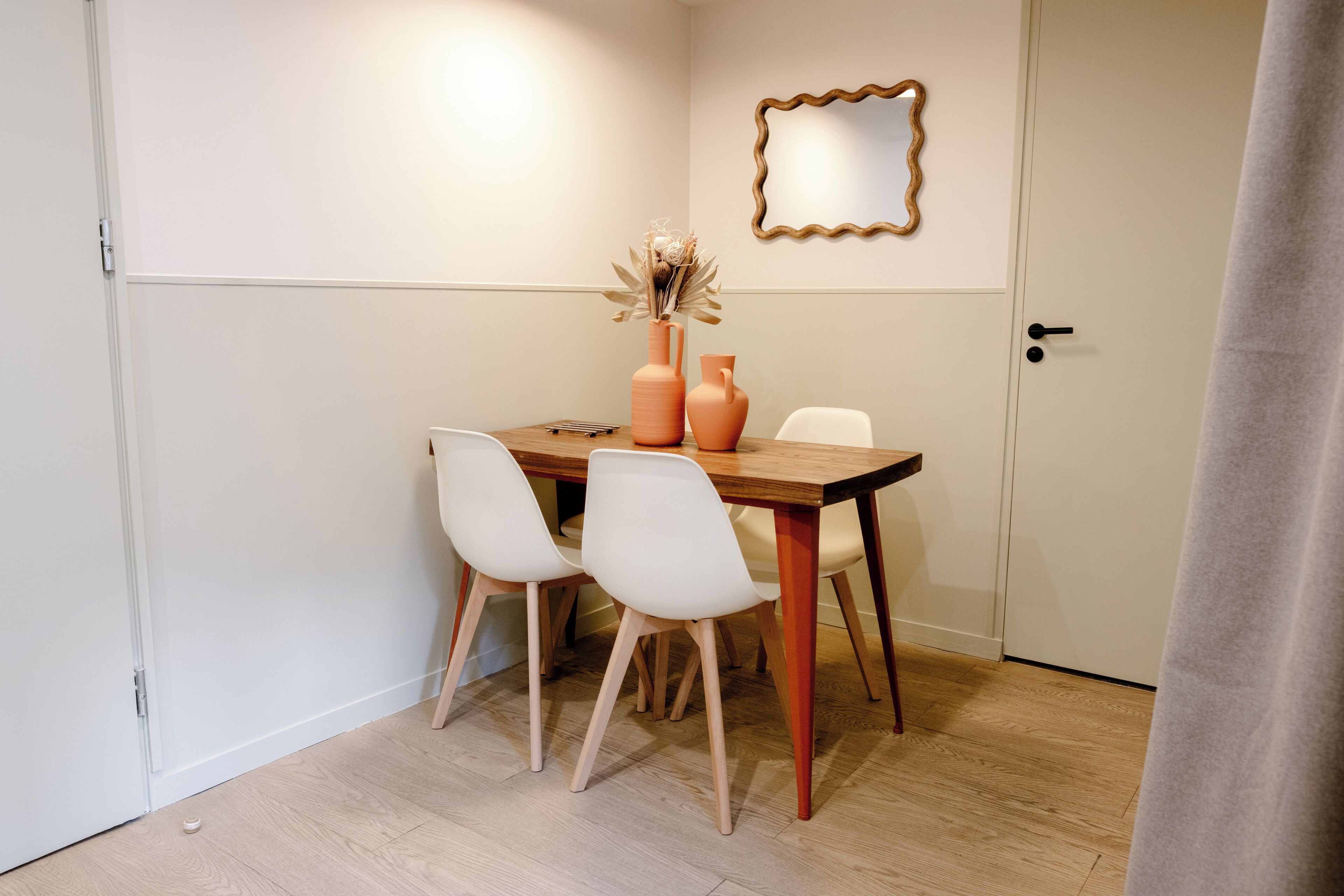 Een eetruimte met een houten tafel, vier witte stoelen, twee vazen met gedroogde bloemen en een spiegel aan de muur.