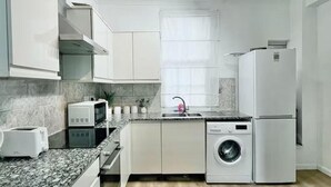 A modern kitchen with white cabinets, a granite countertop, a built-in washing machine, and a window with a white curtain. - Comfortable 3 Bed Flat in King Cross (London)