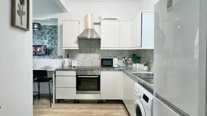 A modern kitchen with white cabinets, a stainless steel range hood, a microwave, an oven, a washing machine, and a trash bin. - Comfortable 3 Bed Flat in King Cross (London)