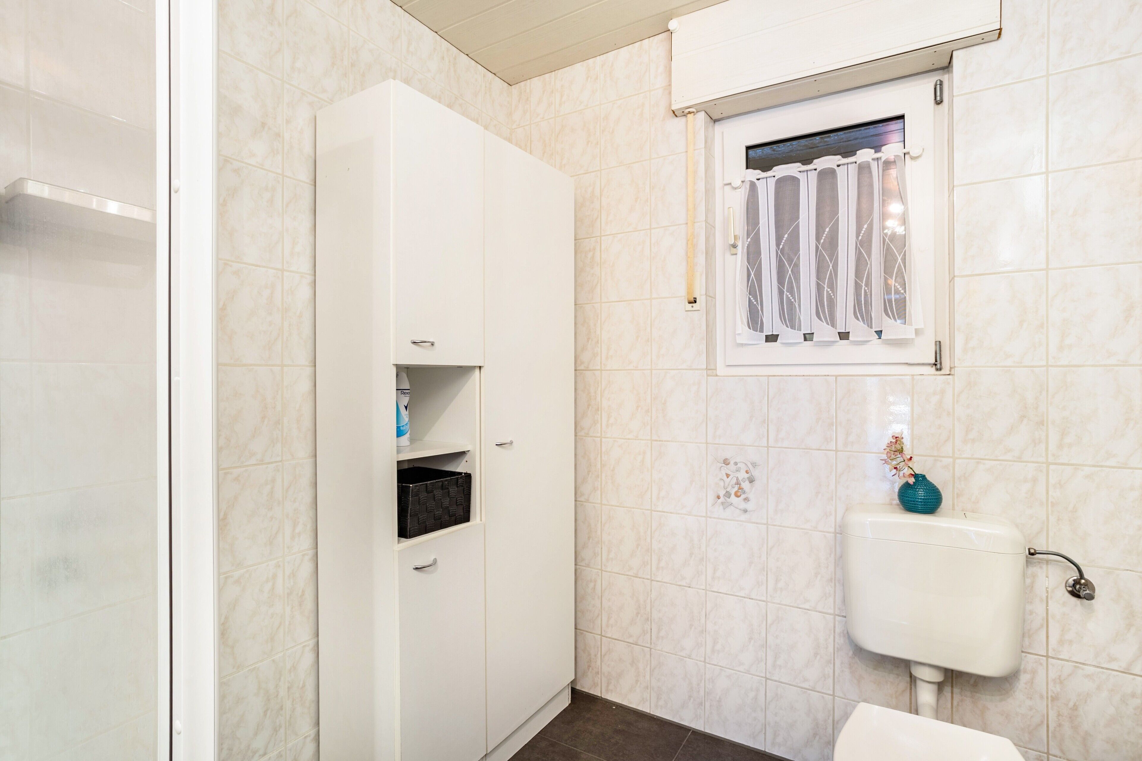 A bathroom with a toilet, a window with a curtain, a cabinet, and a tiled wall.