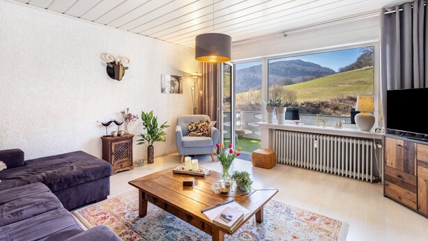 Un salon moderne avec un canapé d’angle, une table basse en bois, un fauteuil, un téléviseur et une grande fenêtre donnant sur un paysage.