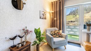 Un salon moderne avec un fauteuil gris, une tête d’animal fixée au mur, un lampadaire et une grande fenêtre donnant sur un paysage verdoyant.