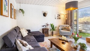 Un salon moderne avec un canapé d’angle, une table basse, une table d’appoint agrémentée d’objets décoratifs, une tête d’animal fixée au mur et une grande fenêtre donnant sur l’extérieur.