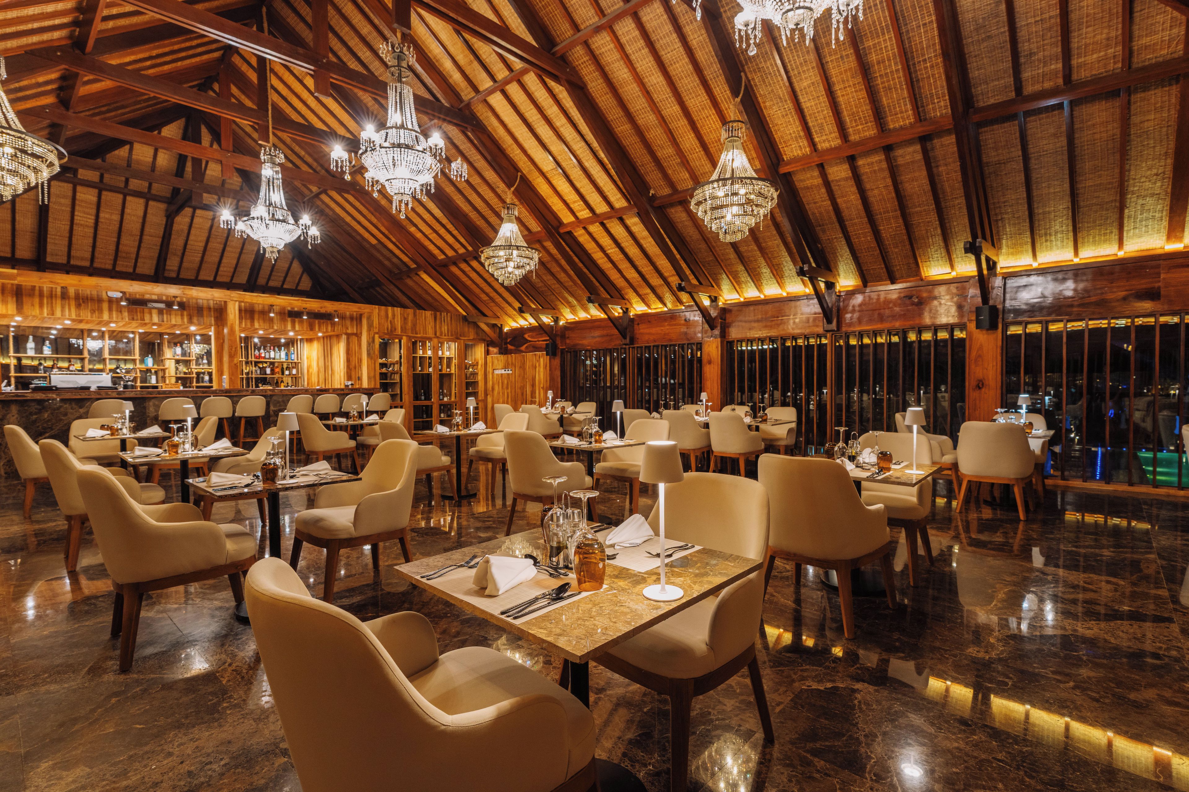Un restaurante espacioso con techos altos, vigas de madera y candelabros. Las mesas están dispuestas con manteles blancos y sillas.