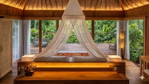 Habitación con techo de madera y cama con dosel. Se observa un banco de madera y una mesita con botellas.