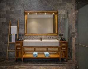 A bathroom with a stone wall, a wooden vanity with a large mirror, and two towels on the floor.