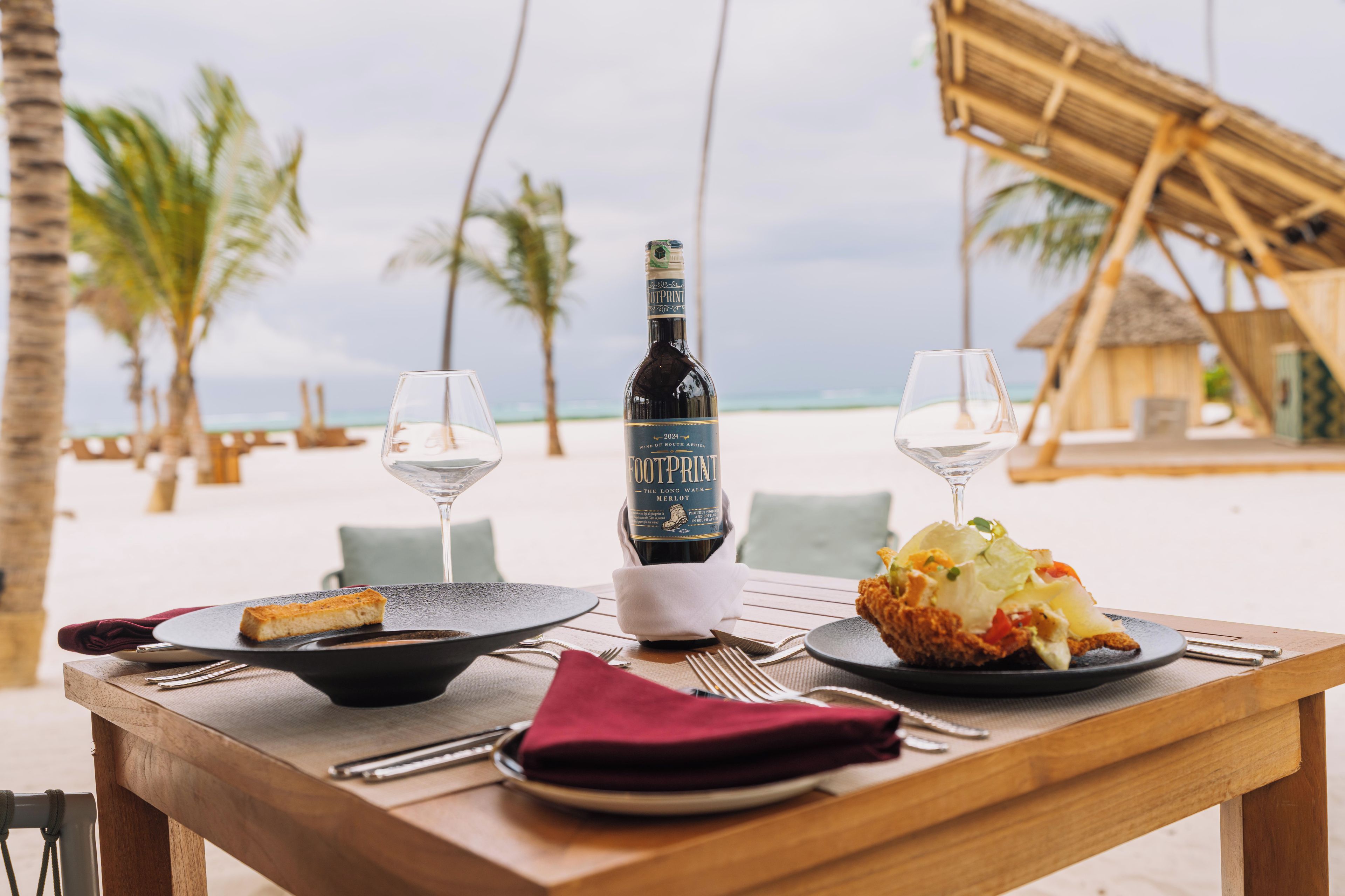 A table set for two with a bottle of wine, two glasses, a plate with food, and a napkin on a wooden table on a beach.