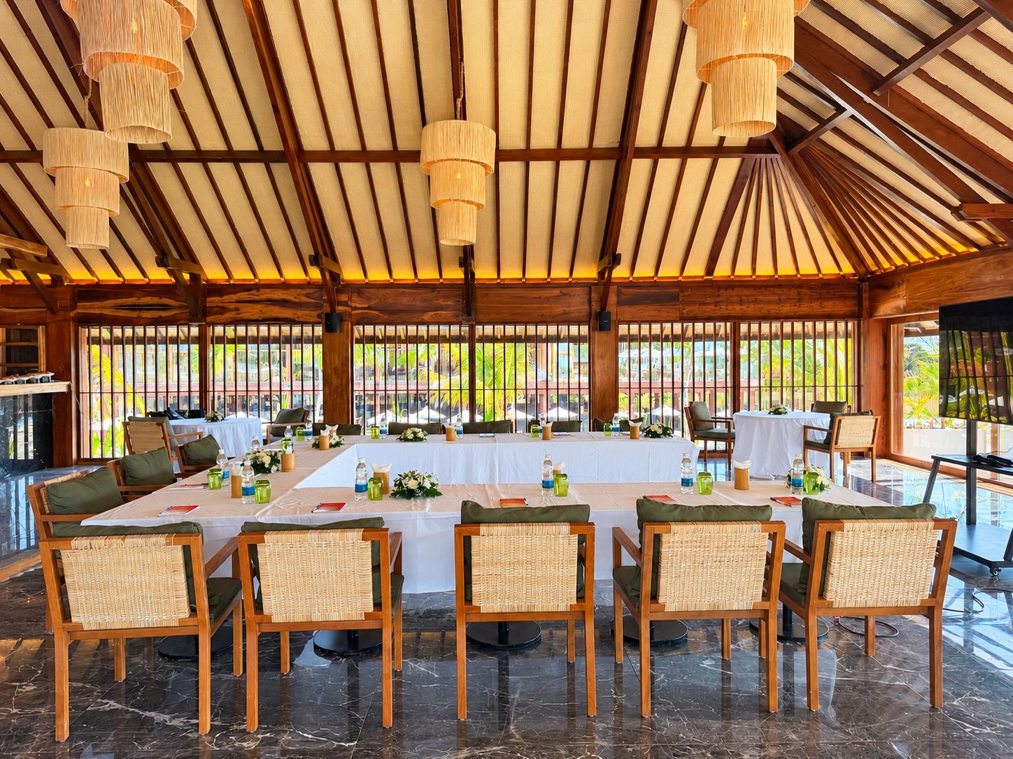 Un comedor interior con una mesa larga dispuesta para una comida, rodeada de sillas de mimbre, bajo un techo de madera con lámparas colgantes.