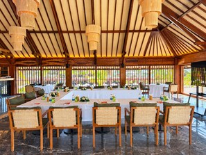 Un espace de restauration intérieur avec une longue table dressée pour un repas, entourée de chaises en osier, sous un plafond en bois agrémenté de lampes suspendues.