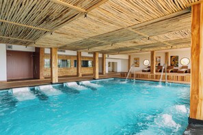 Een groot binnenzwembad met houten balken in het plafond, een whirlpool en een zithoek met kussens.