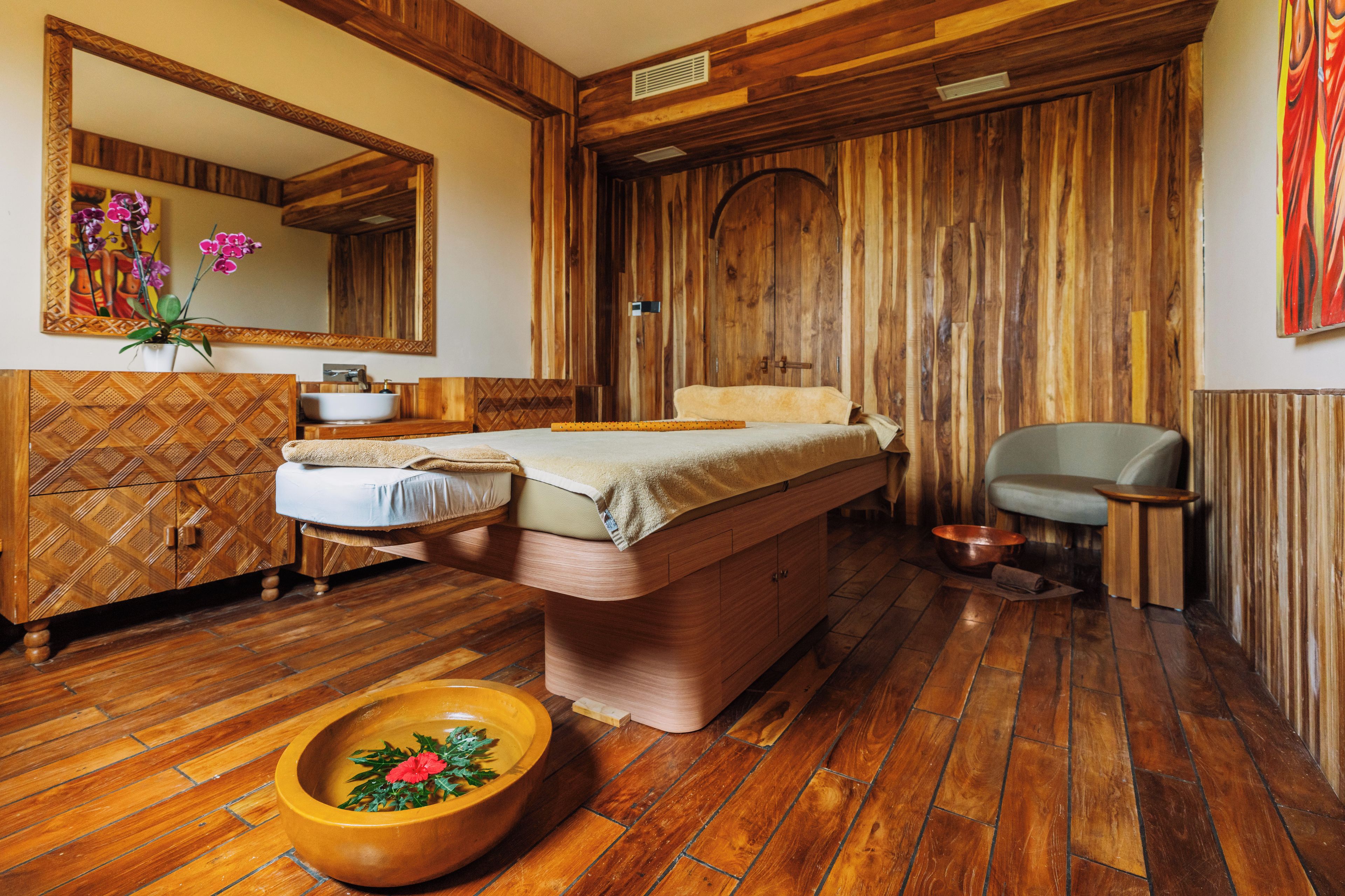 Una habitación con suelo de madera, una cama, una mesita, una silla, un lavamanos y un espejo grande.