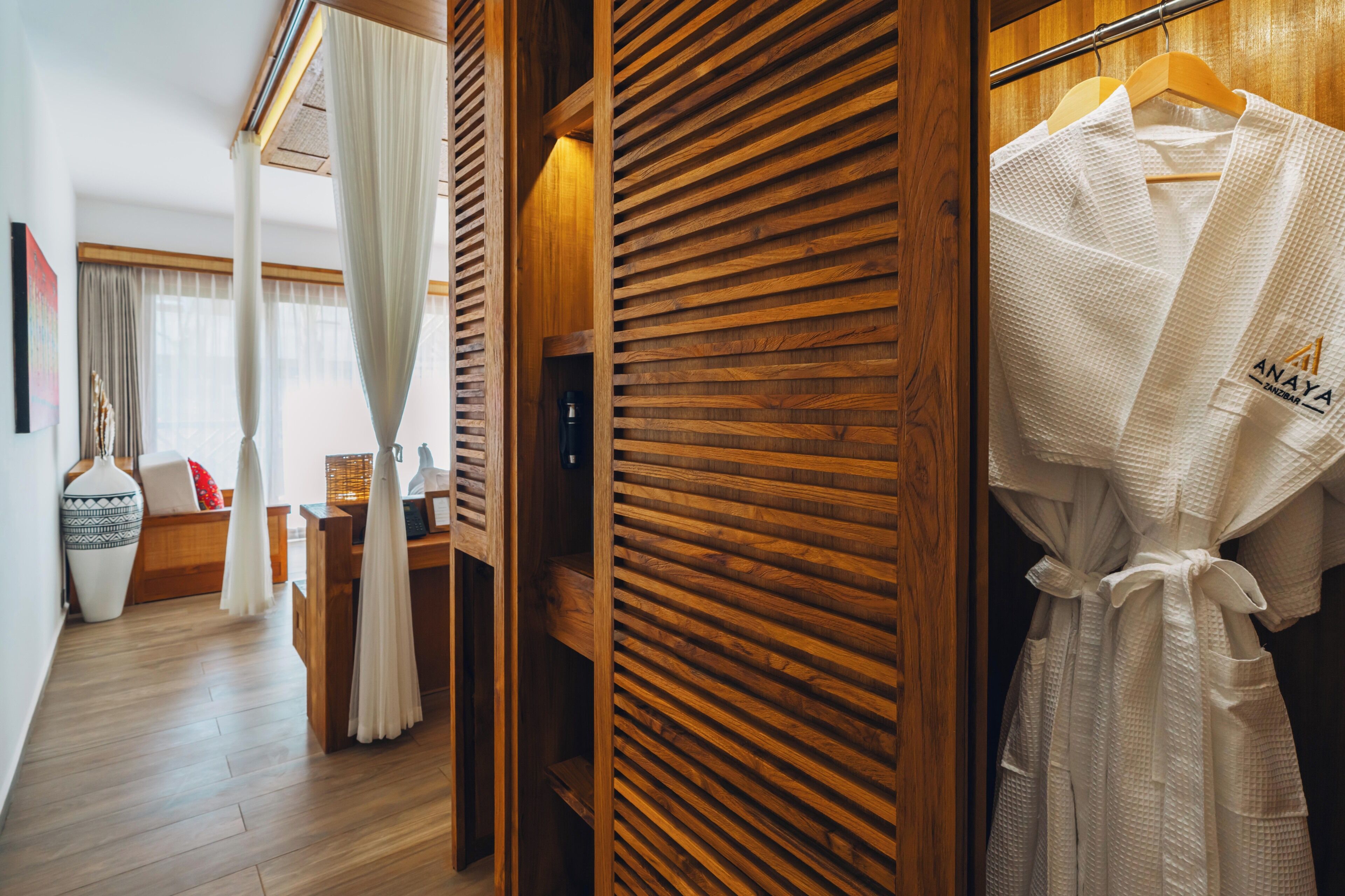 Quarto de hotel com guarda-roupa de madeira, um roupão branco pendurado em um cabide de madeira, chão de madeira e um vaso grande sobre uma mesa lateral.