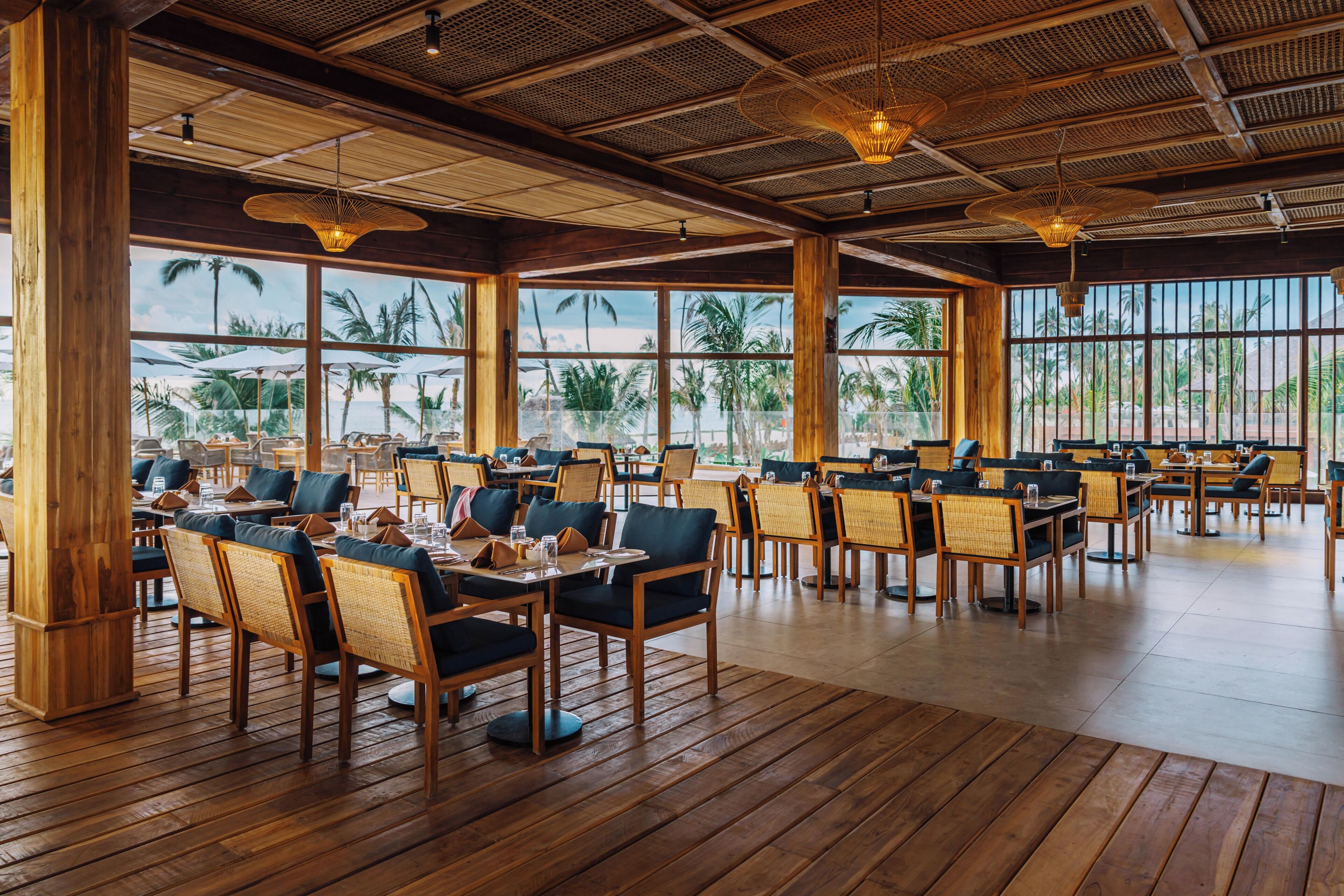 A spacious indoor dining area with wooden floors and tables, large windows offering views of palm trees and the sea.