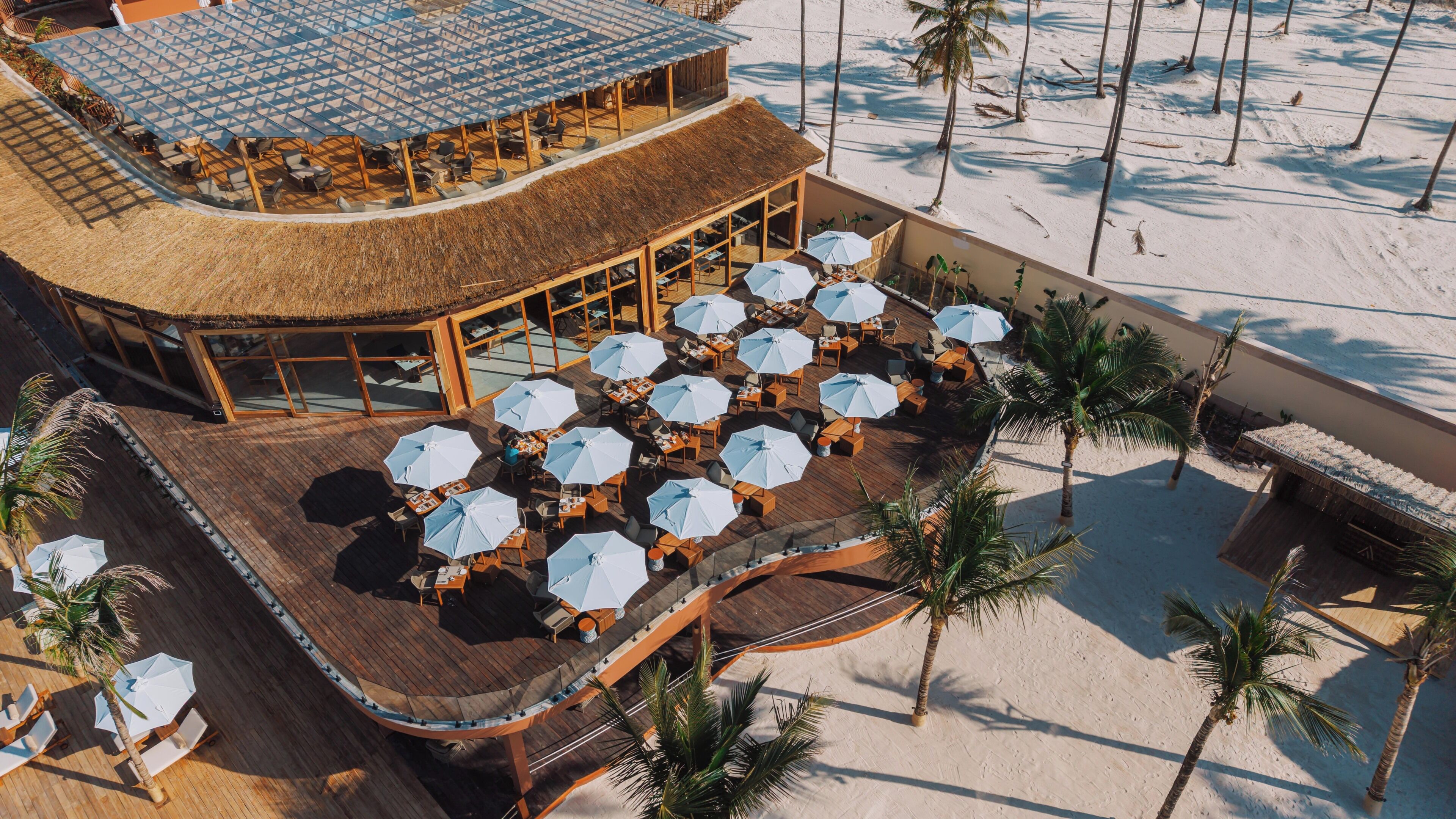 Un resort frente a la playa con asientos exteriores y sombrillas, un edificio de techo de paja y palmeras.