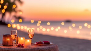 Une table dressée avec des bougies, des verres et une assiette de nourriture, sur une plage au coucher du soleil.