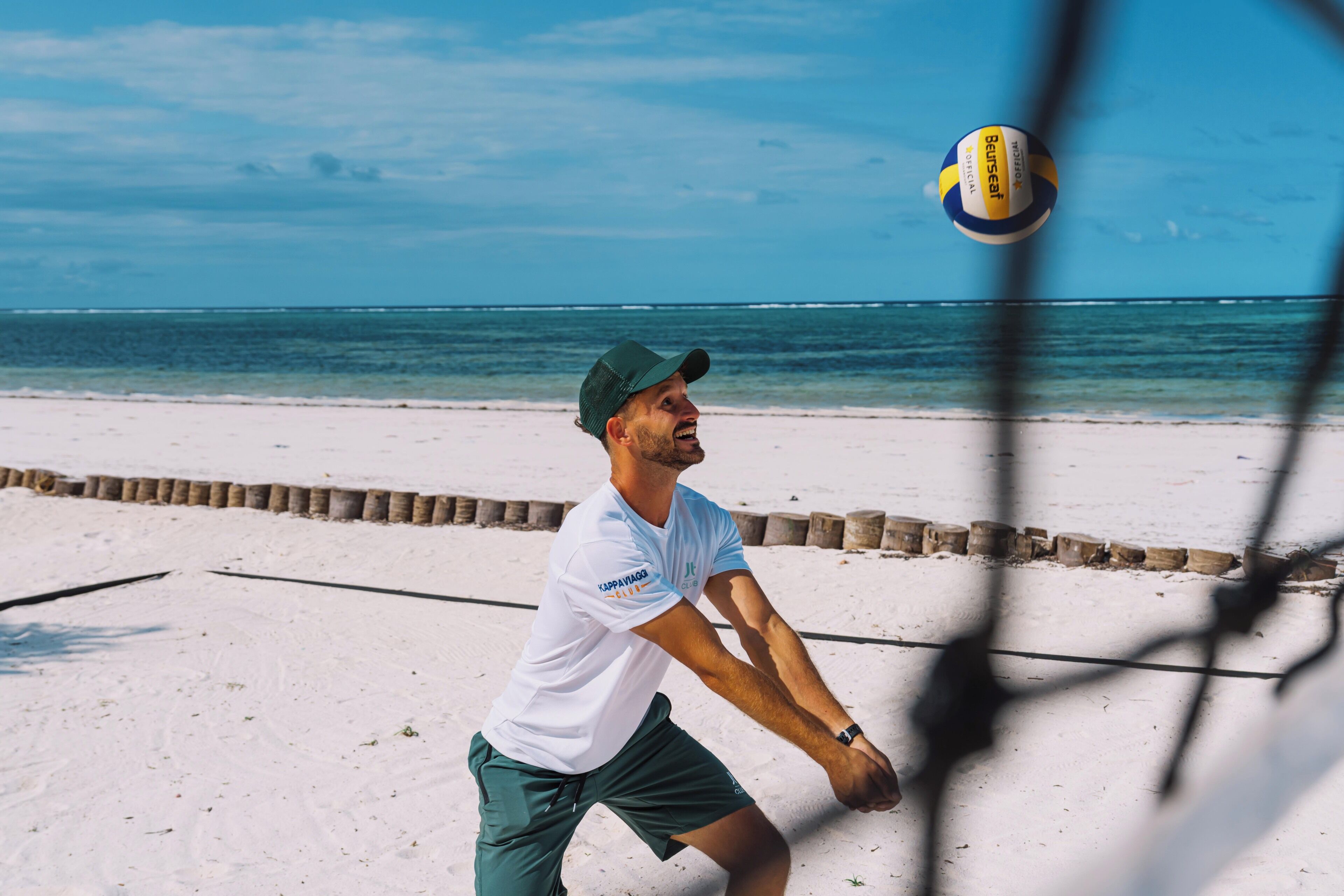 Um jogo de voleibol de praia, com uma rede em primeiro plano e uma praia arenosa com uma barreira de madeira ao fundo.