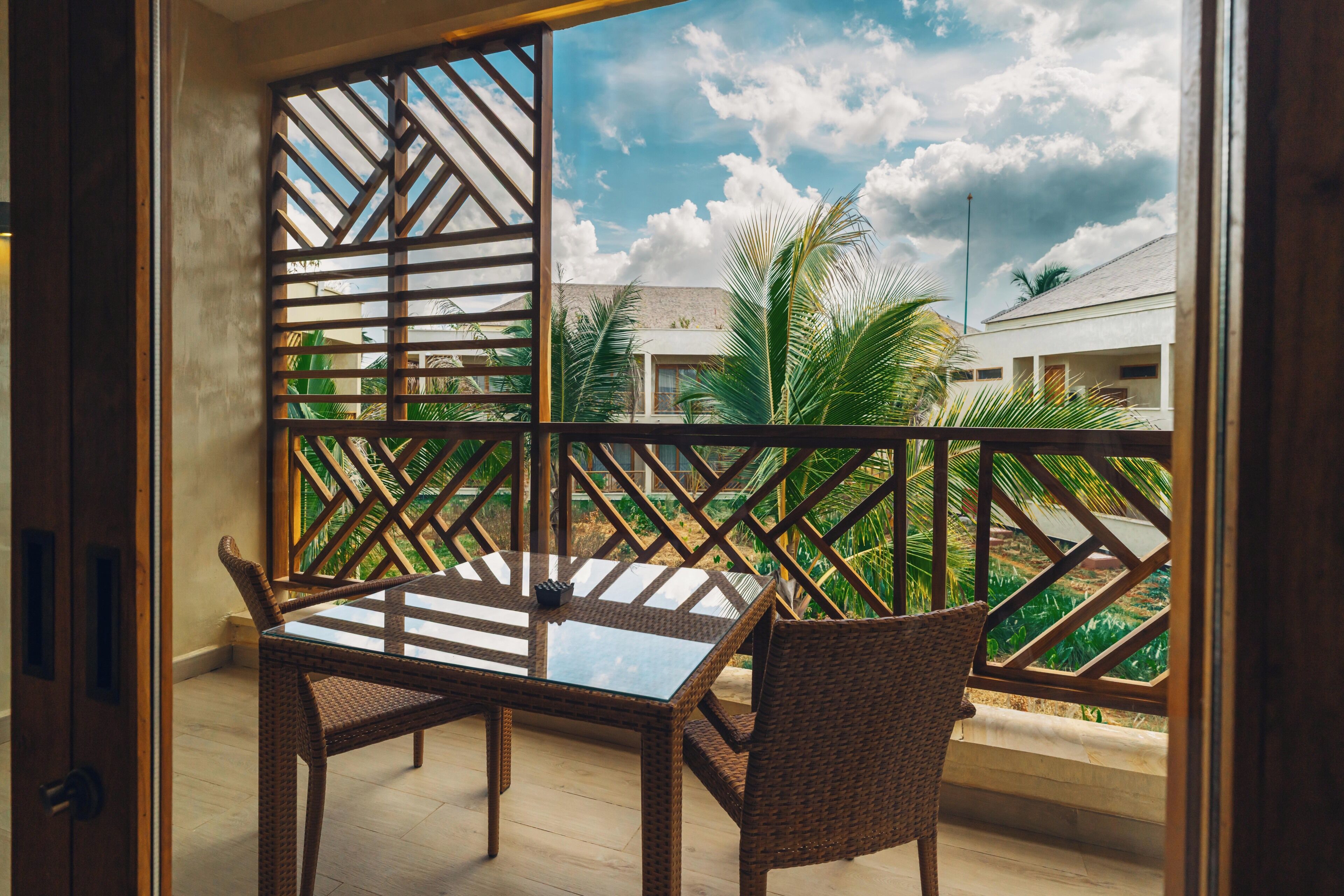 Uma varanda com mesa de vidro e cadeiras de vime, com vista para uma área residencial com palmeiras e casas.