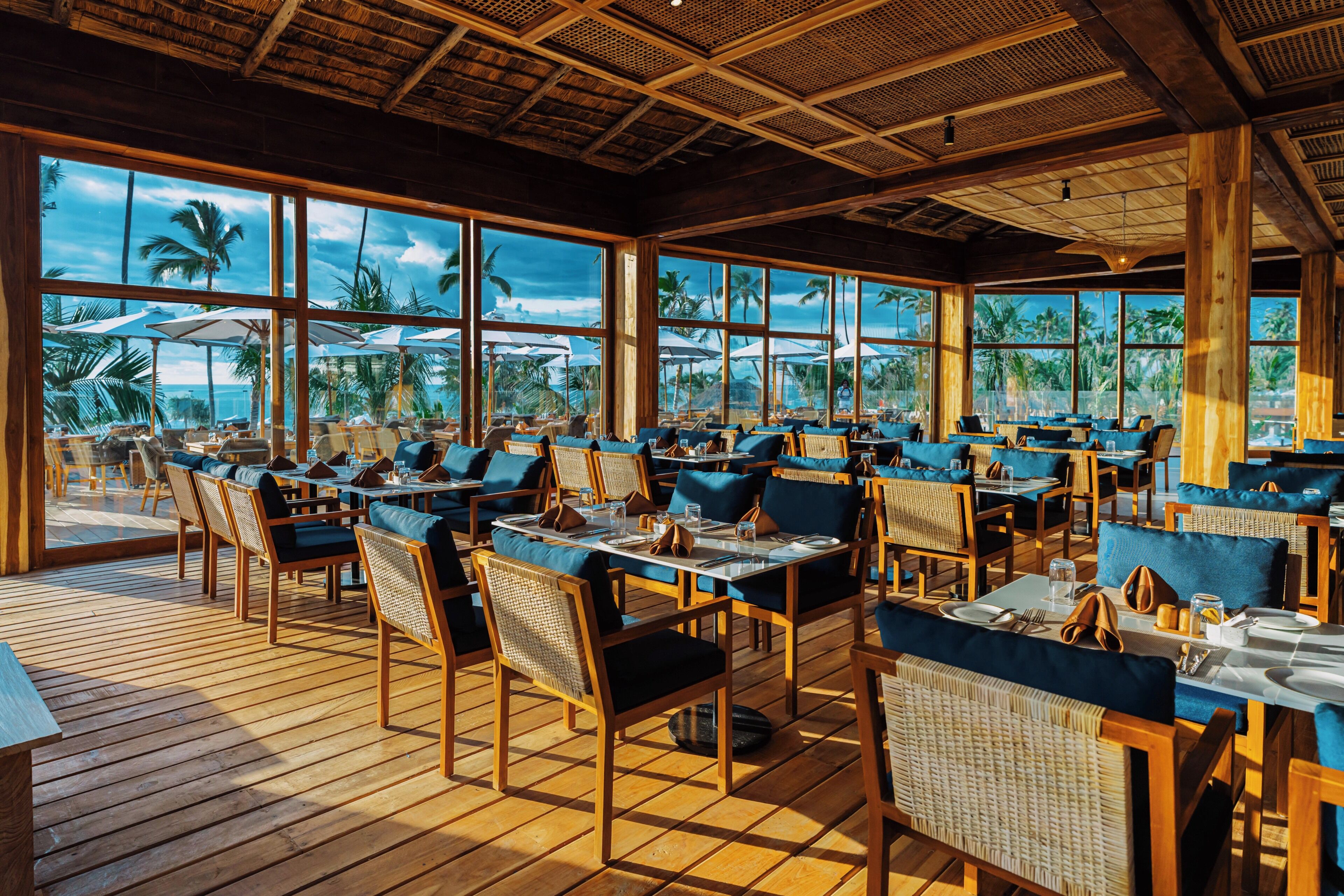A spacious outdoor dining area with wooden tables and chairs, large windows offering views of palm trees and the ocean.