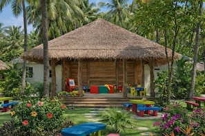 A thatched-roof building with colorful play equipment and benches in a tropical garden setting.