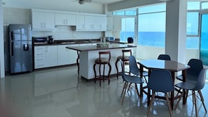 Una cocina moderna con armarios blancos, una isla central con taburetes y vista al océano.