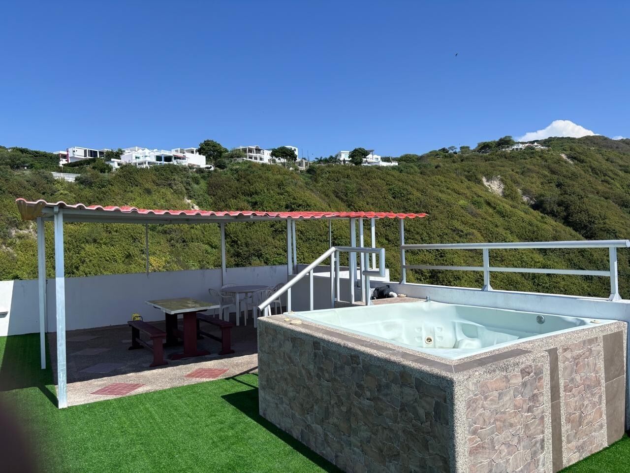 A rooftop terrace with a hot tub, outdoor seating, and a view of a hillside with buildings.