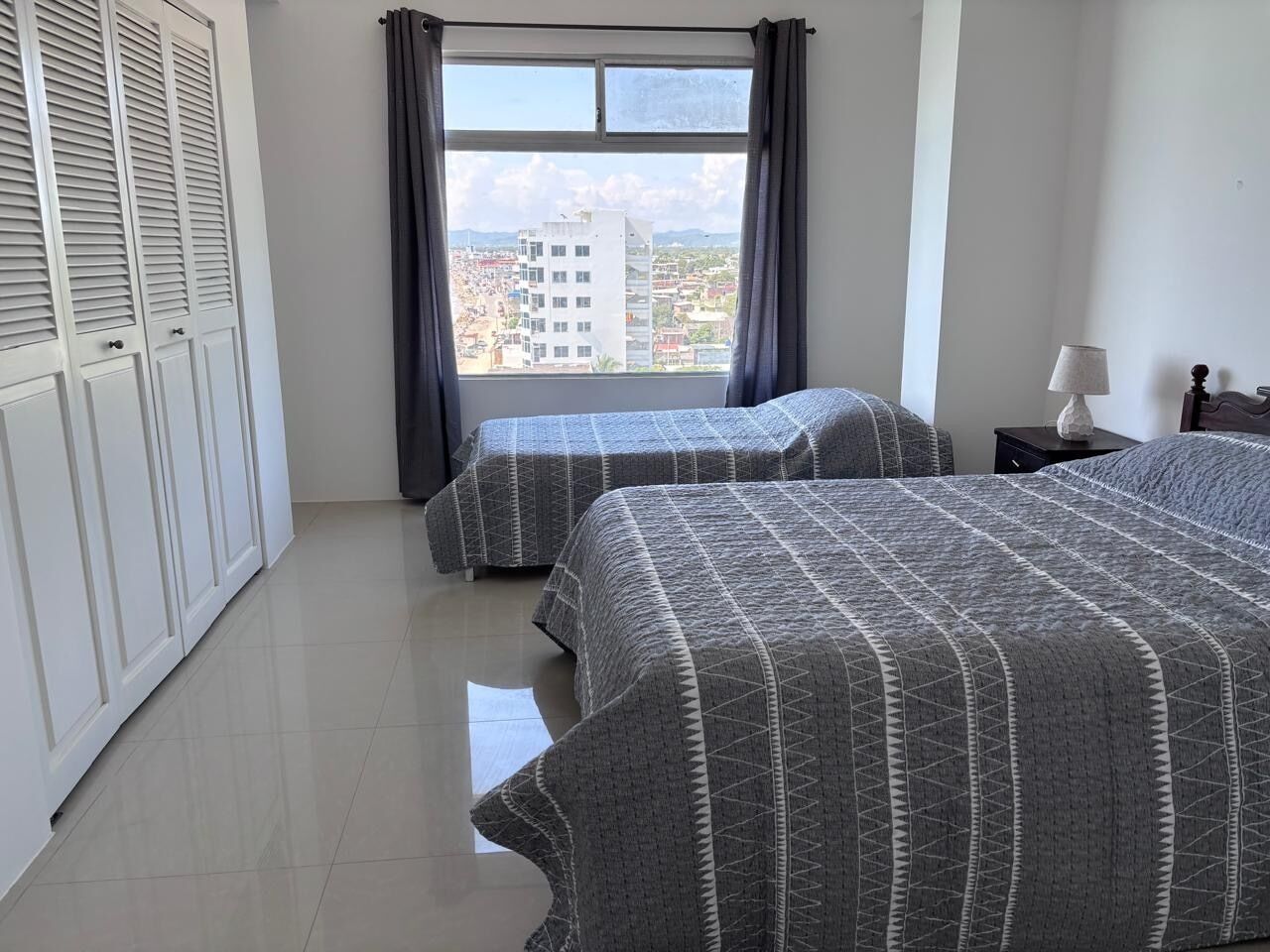 Camera da letto con due letti, una finestra con tende, un comodino e vista sugli edifici esterni.