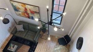 Una sala de estar moderna con un sofá verde, una mesa de centro y una mesa redonda. Hay un cuadro abstracto grande en la pared y una ventana con vistas a un edificio de ladrillo.