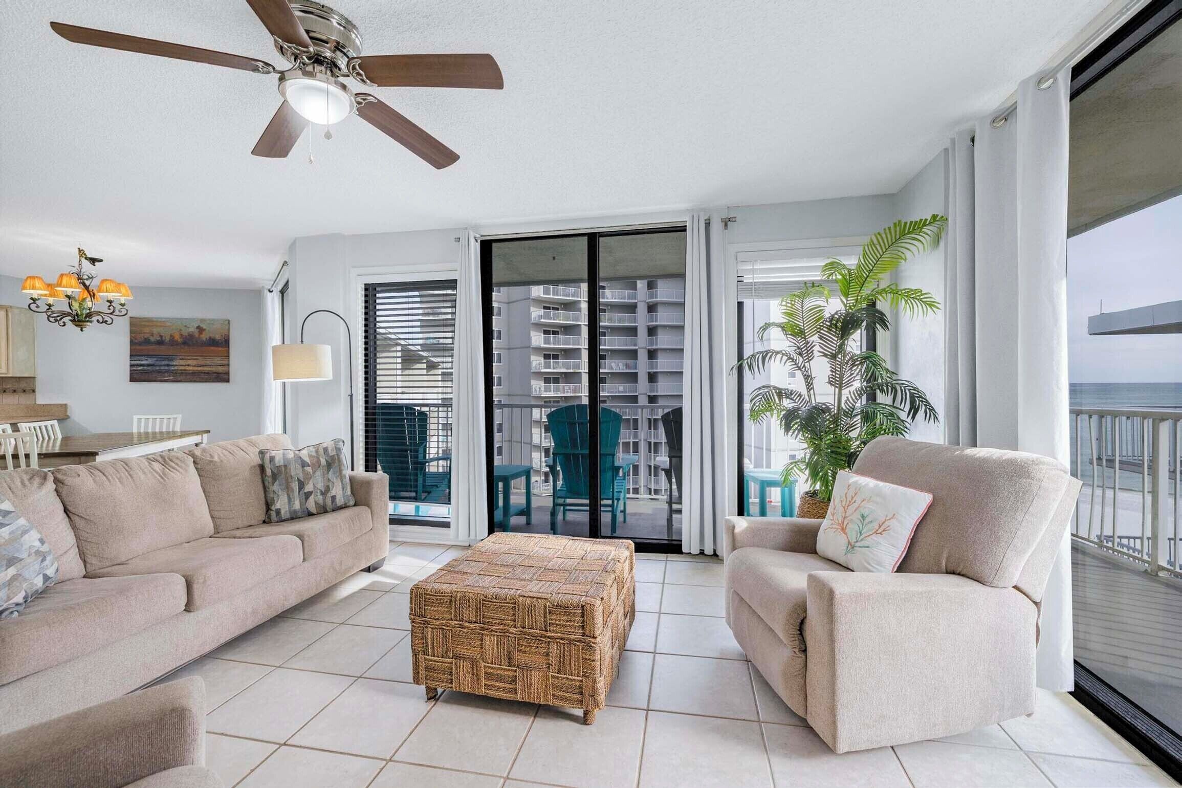 A modern living room with a ceiling fan, a sofa, a chair, a wicker ottoman, and a view of a balcony with outdoor furniture and a plant.
